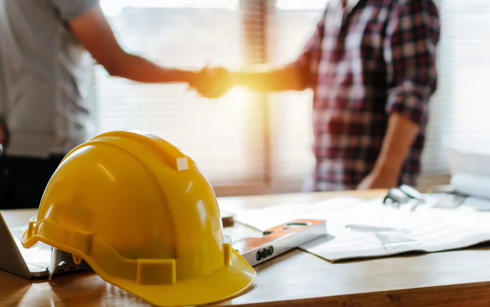 yellow safety helmet on workplace desk with construction worker team hands shaking greeting start up plan new project contract in office center at construction site, partnership and contractor concept